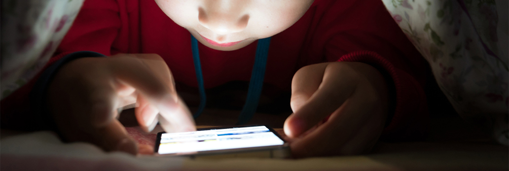 A child looks at a mobile device at night beneath a duvet